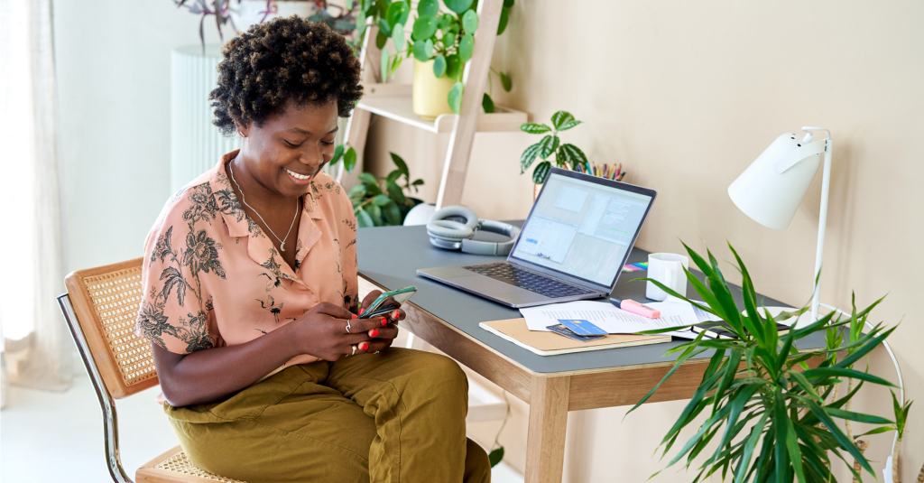 A person smiling while looking at their phone and sitting at a desk with a laptop on it