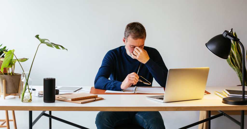 Man sitting at desk, pinching bridge of his nose while sitting in front of a laptop