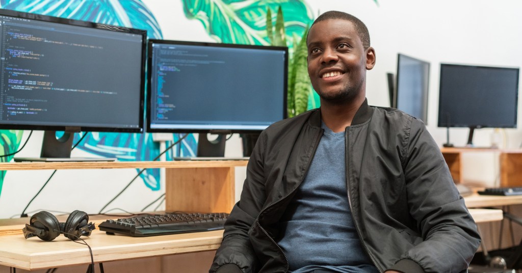 A person sitting at chair in front of a desk with two monitors while turned away and smiling up