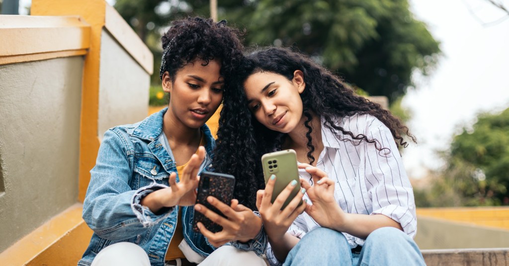 Two people sitting while on their cellphones. One person is sharing what they are looking at with the other person