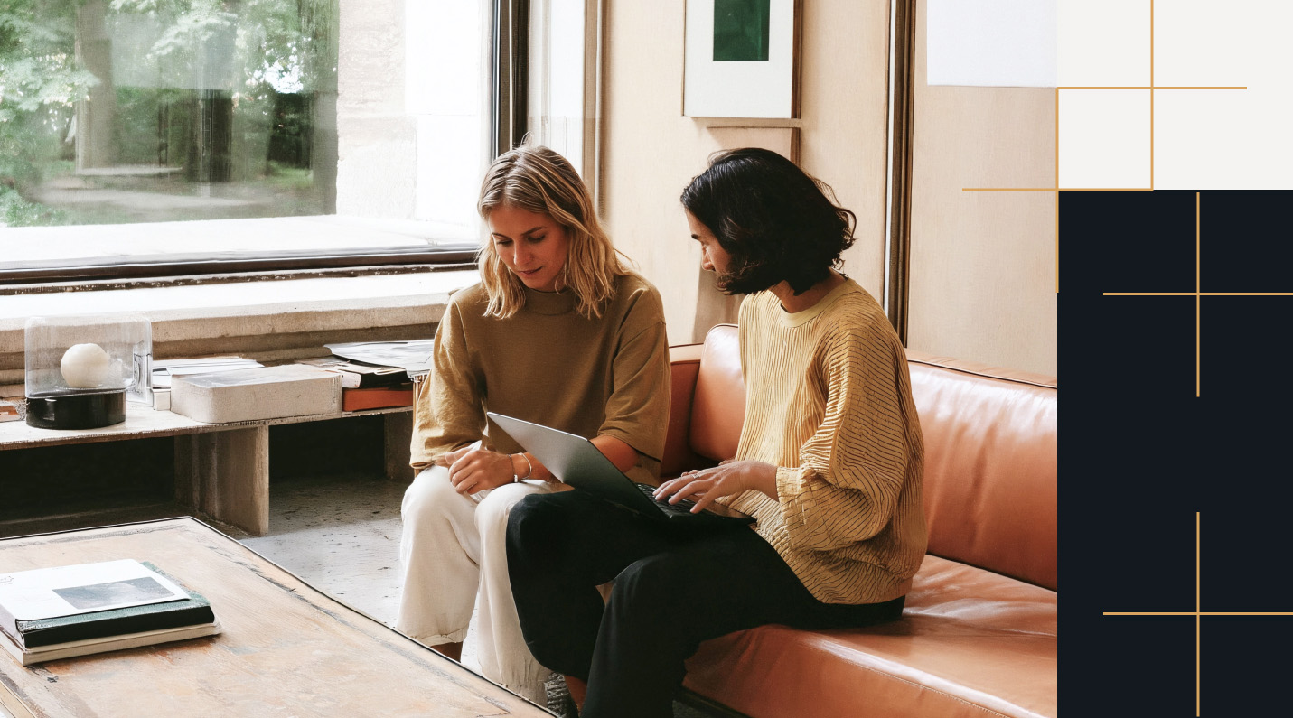 Two women collaborating in a modern office with natural light. One woman with dark hair in a yellow textured sweater points at a laptop screen, while the other woman with blonde hair and a tan sweater looks on attentively. They sit on a leather sofa next to a large window, with minimalist decor and books around them.